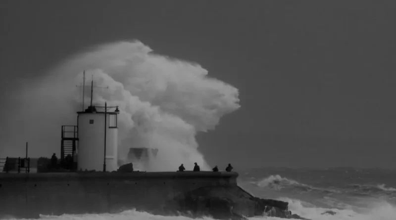 A huge wave crashing over a lighthouse and sea wall in black and white